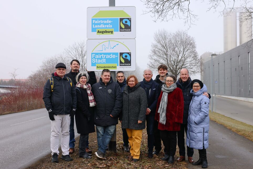 Eine Gruppe von Personen steht lächelnd an einem Straßenrand neben Schildern mit der Aufschrift Fairtrade-Landkreis Augsburg und Fairtrade-Stadt Schwabmünchen.