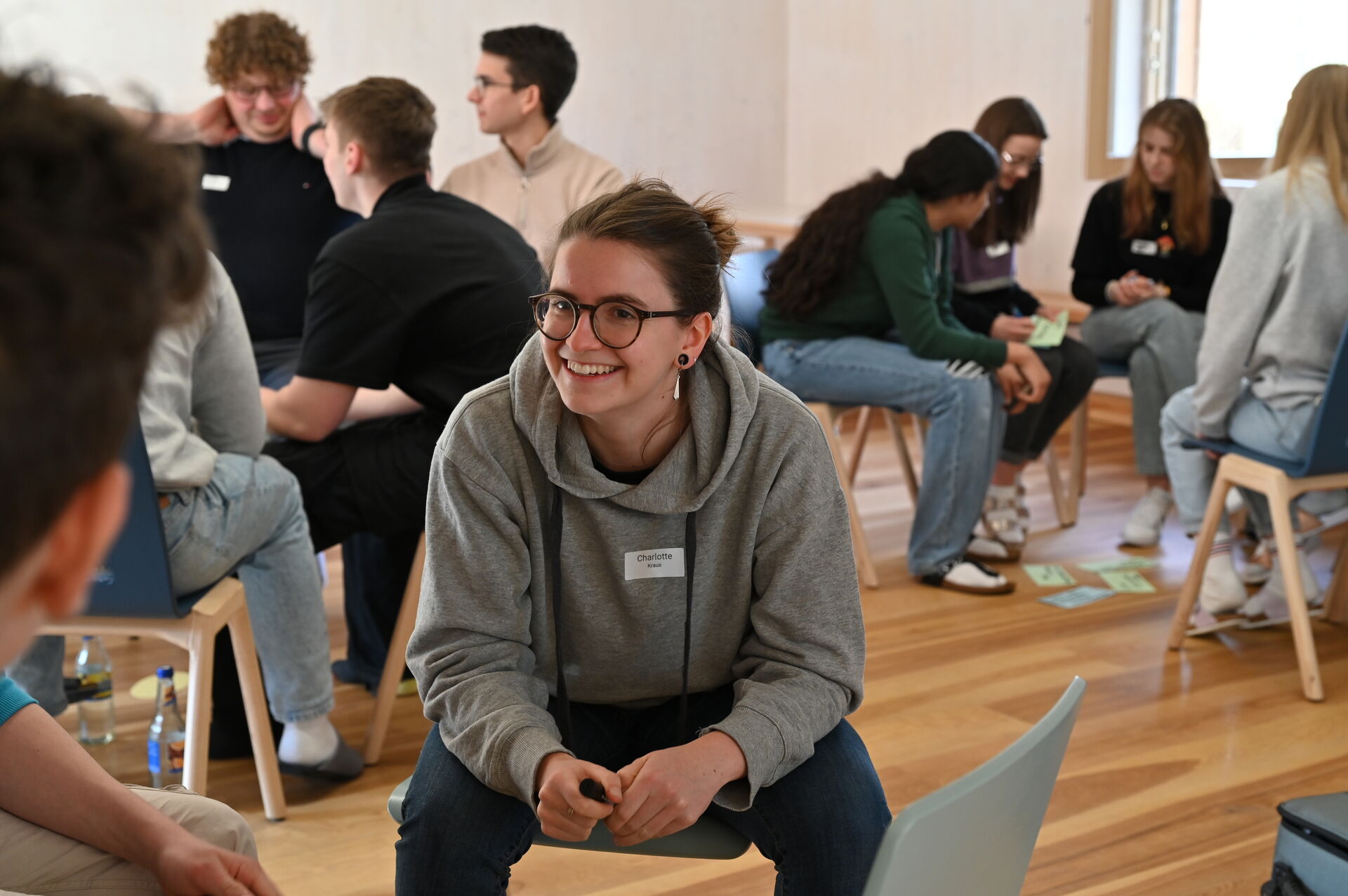 Eine Gruppe junger Erwachsener sitzt in einem kleinen Kreis und unterhält sich in einem hellen Raum. Eine lächelnde Person mit Brille und grauem Kapuzenpulli sitzt im Vordergrund und schaut jemanden außerhalb des Bildes an.