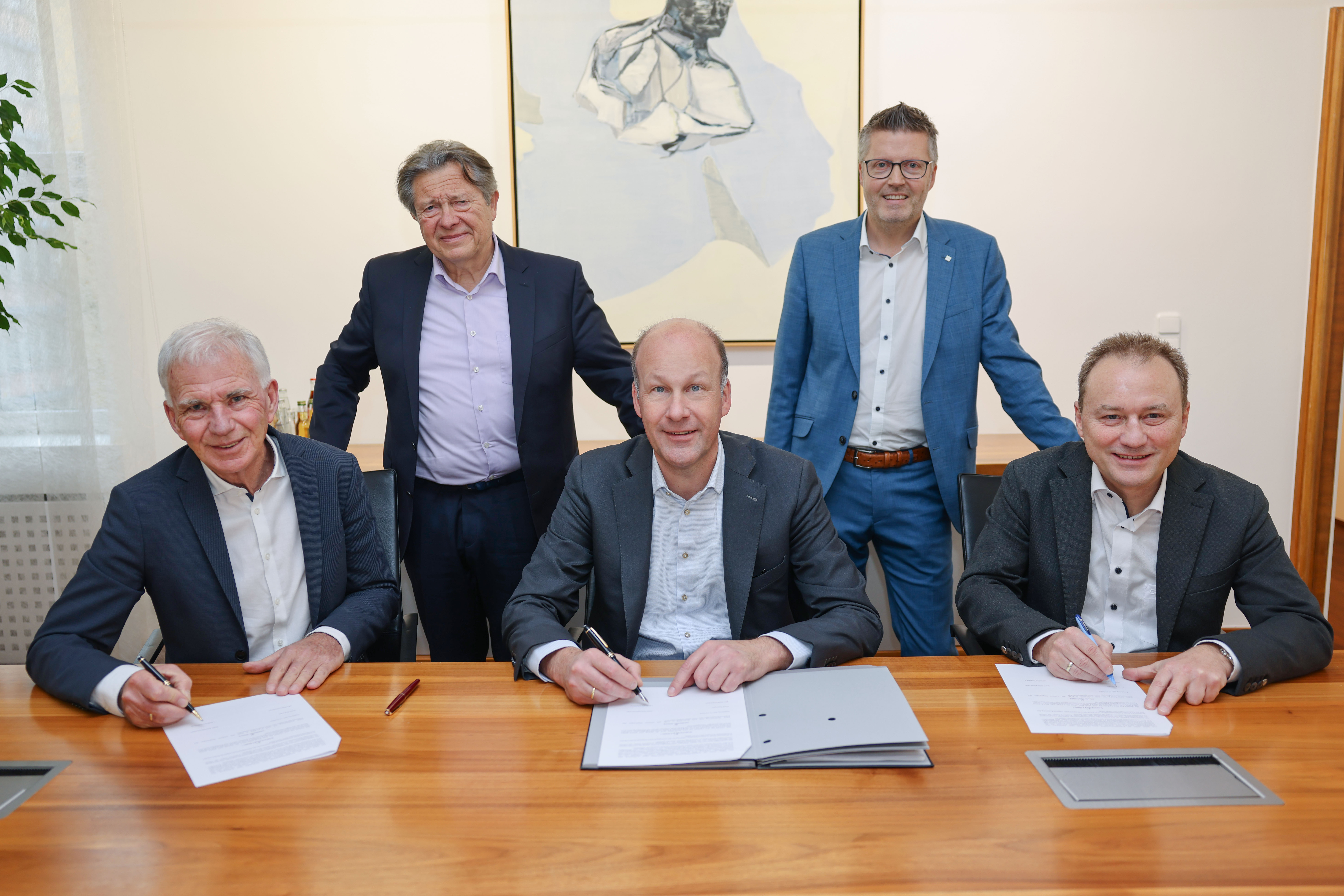 Fünf Männer in Geschäftskleidung, drei von ihnen sitzen und unterschreiben Dokumente an einem Holztisch, zwei stehen hinter ihnen. An der Wand im Hintergrund hängt ein Gemälde. Die Umgebung scheint ein formelles Büro oder ein Besprechungszimmer zu sein.